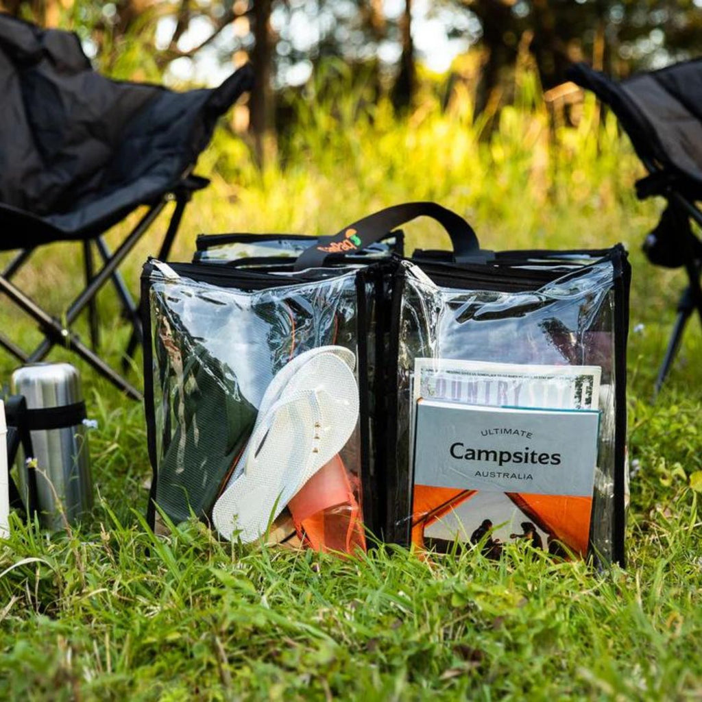 Large black CamPaq clear bag filled with books shoes and camping items sitting on grass beside camp chairs