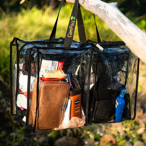 Clear large black trim camping bag with black handles filled with toiletries, hanging  on tree outdoors.